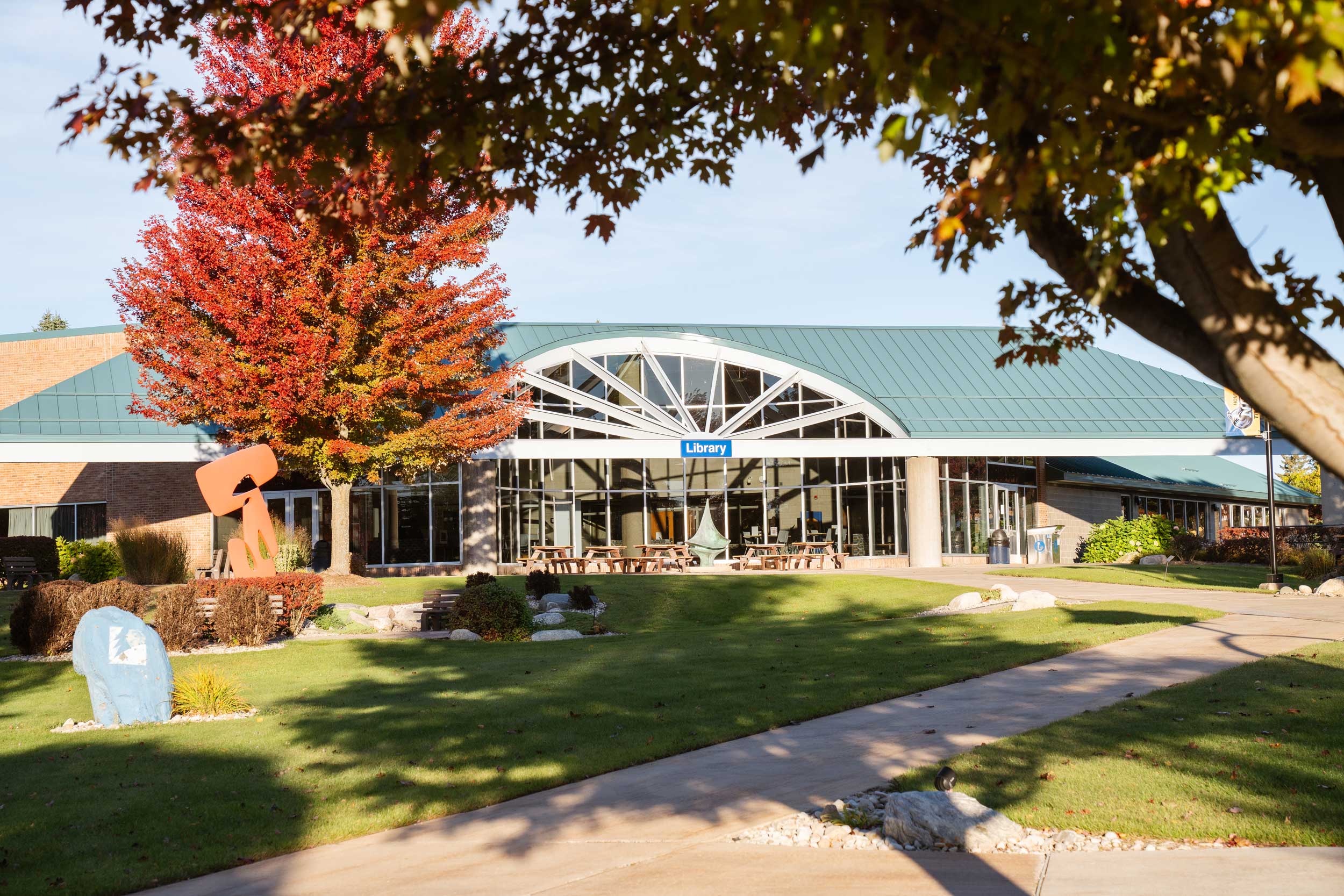 Exterior view of the North Central Michigan College Library on a sunny fall day, with trees in autumn color and campus walkways leading to the building’s main entrance
