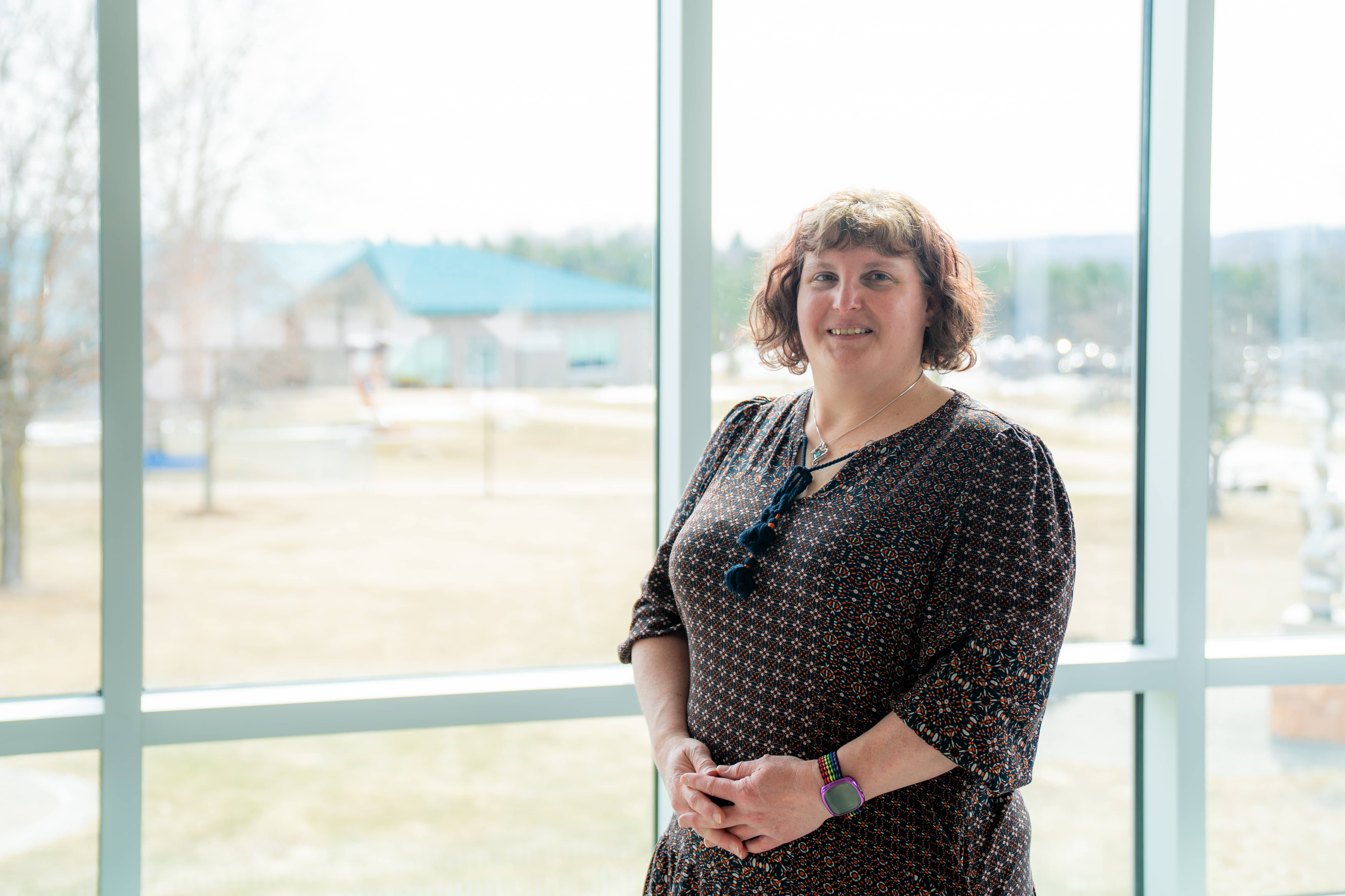 A smiling woman stands in front of a large window at North Central Michigan College, wearing a patterned blouse and a colorful watch. Natural light fills the space, with a blurred outdoor view of the campus in the background.