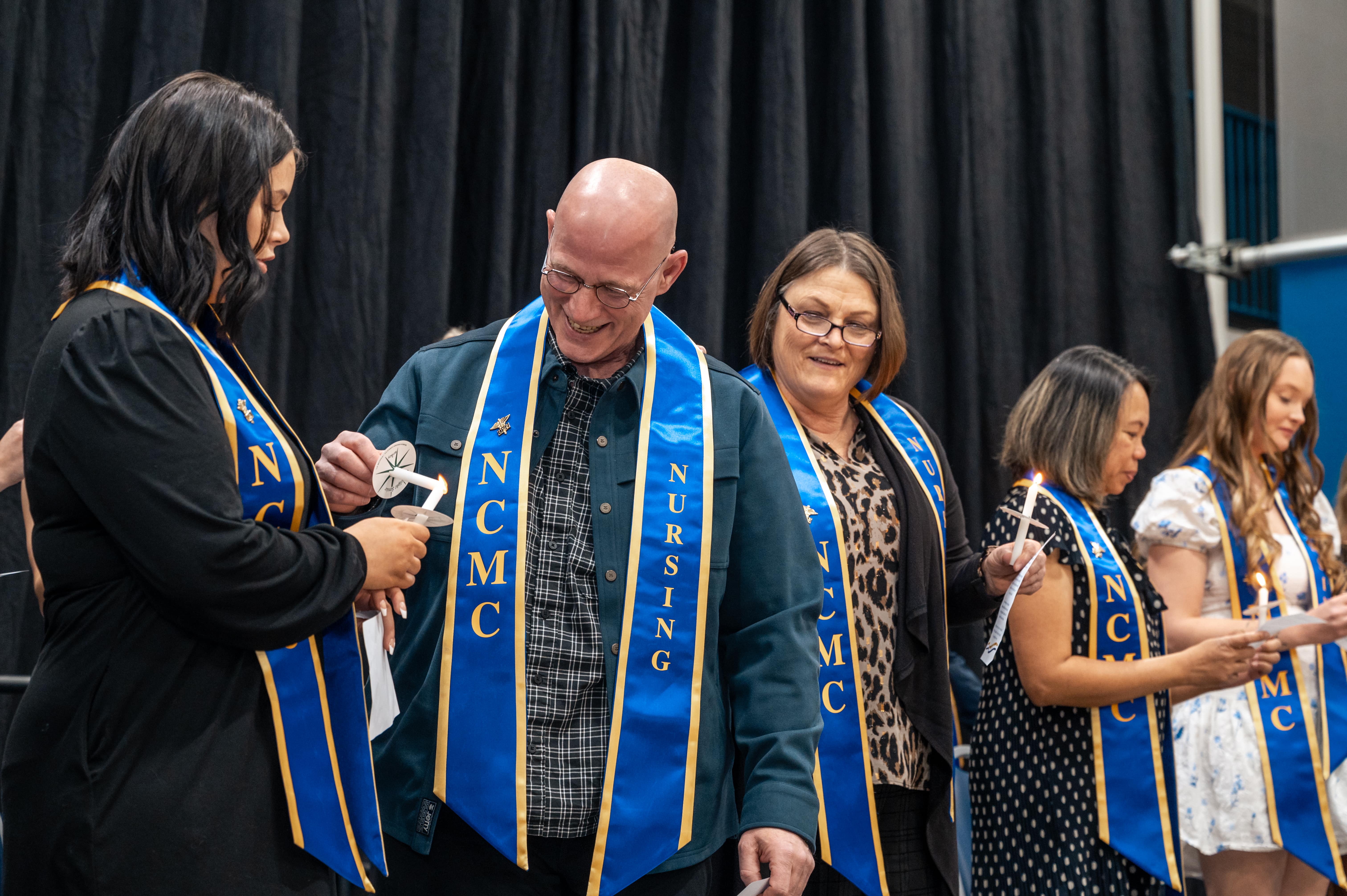 A group of North Central Michigan College Practical Nursing graduates wearing blue and gold NCMC stoles participate in the candle-lighting portion of the pinning ceremony. One graduate lights another’s candle while others stand nearby holding their candles.