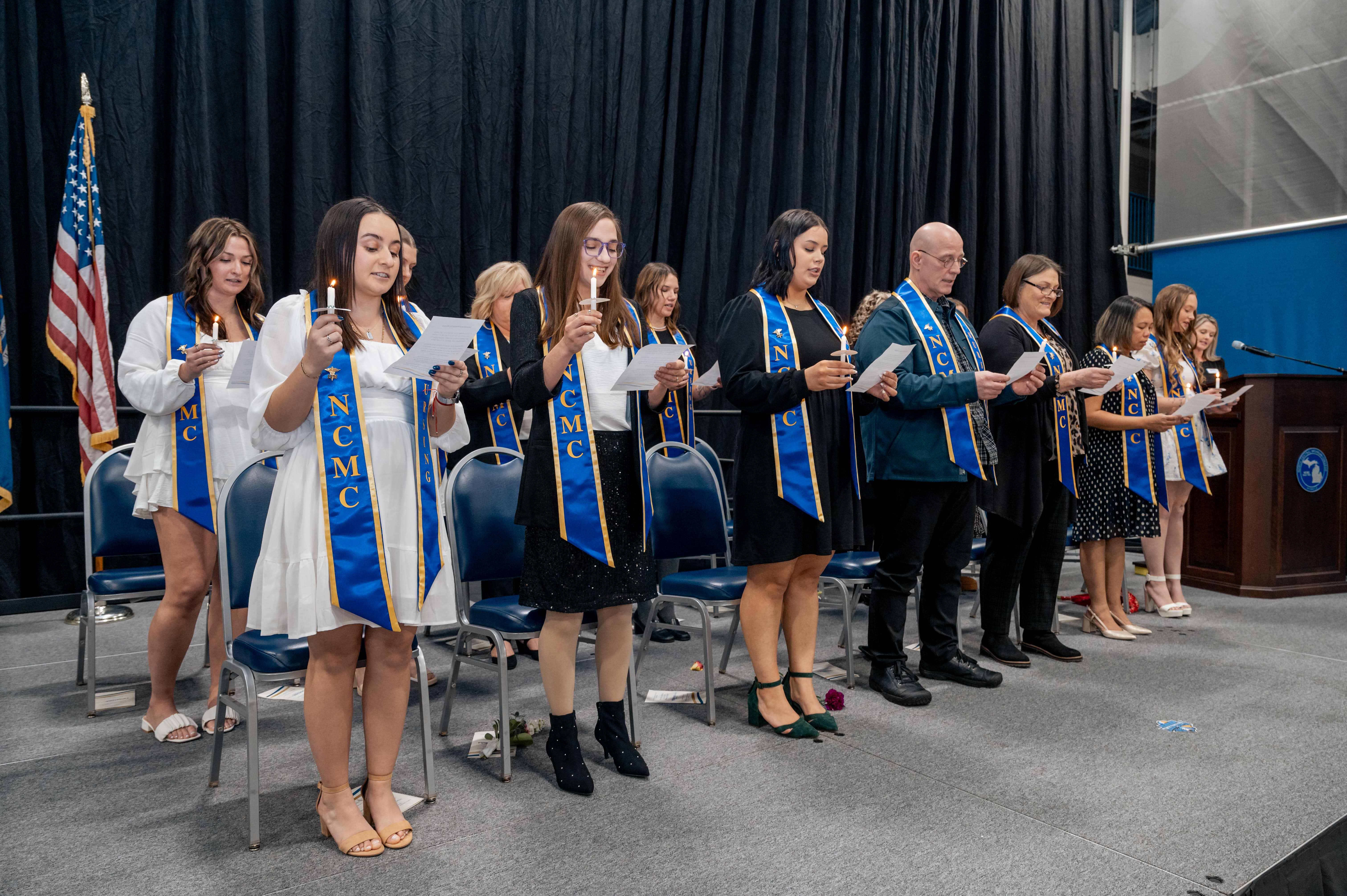 North Central Michigan College Practical Nursing graduates stand two rows on stage, wearing blue and gold NCMC stoles and holding lit candles as they recite the nursing pledge during the pinning ceremony.