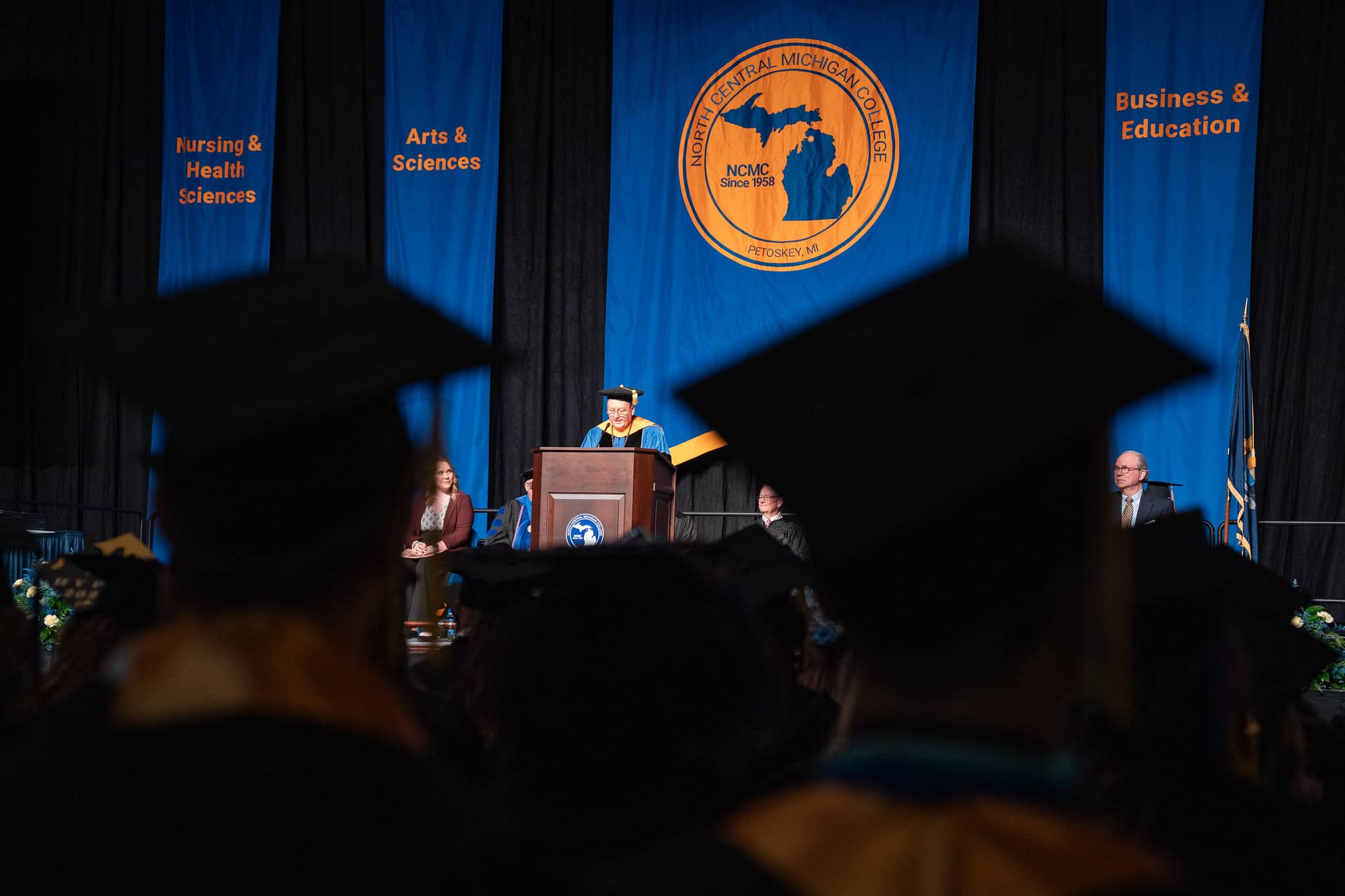 Speaker at podium during North Central Michigan College graduation ceremony, with graduates in caps and gowns in the foreground