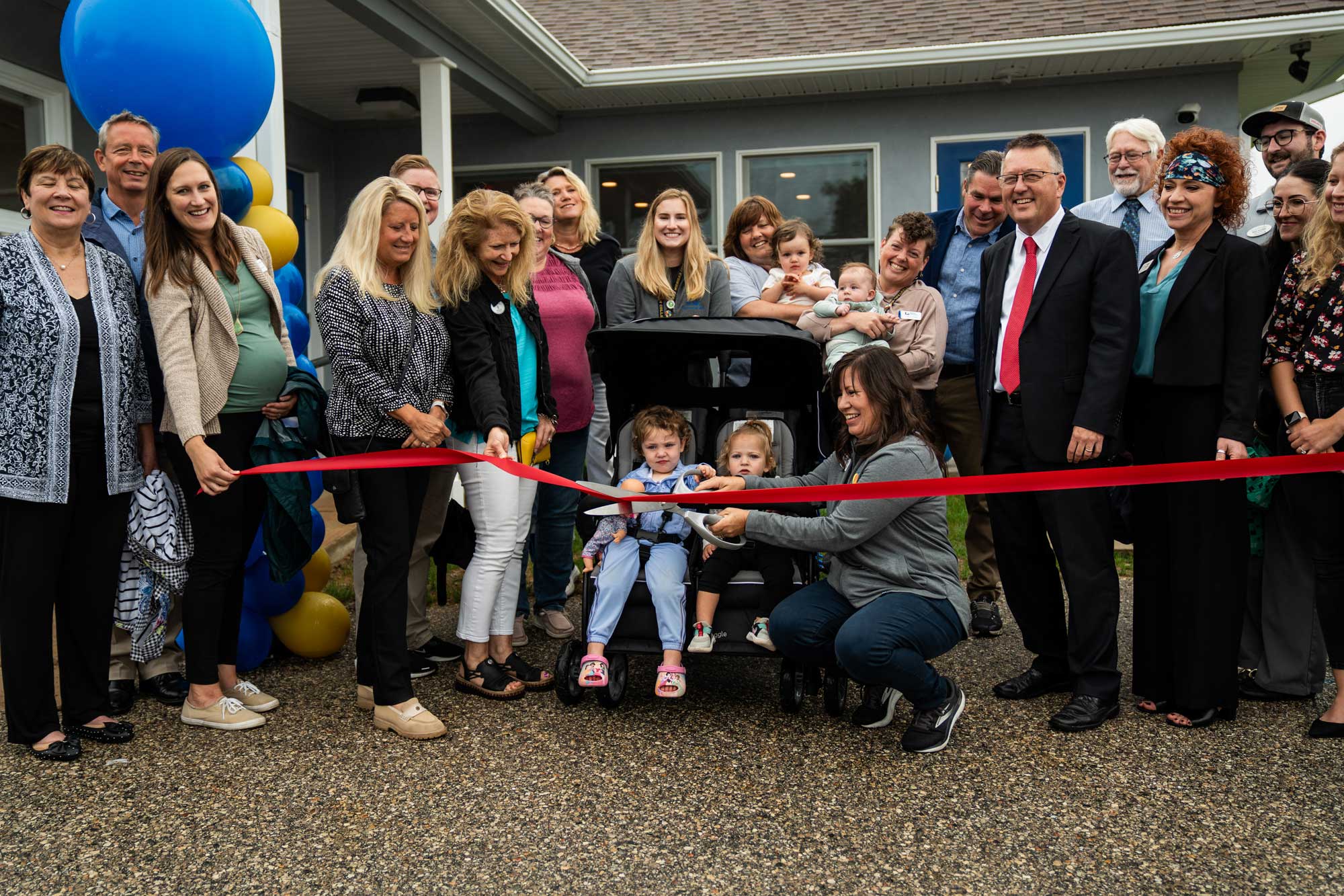 A group of community members, families, and NCMC leaders gathers outside the new Early Learning Center as children help cut a red ribbon during the opening ceremony.