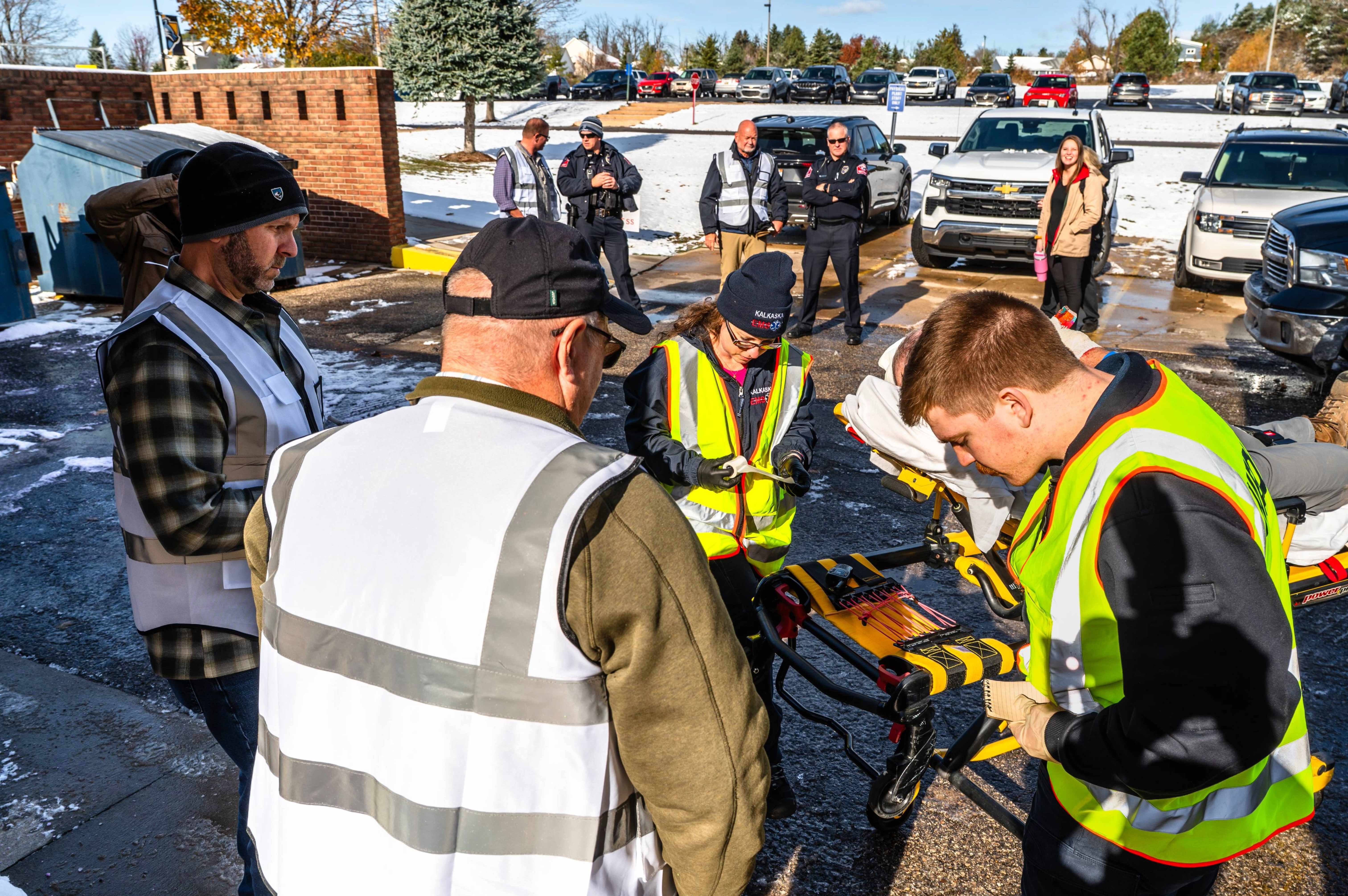 North Central Michigan College EMS students wearing high-visibility safety vests work together to treat and transport a simulated patient on a stretcher during an outdoor emergency response training exercise while instructors and first responders observe.