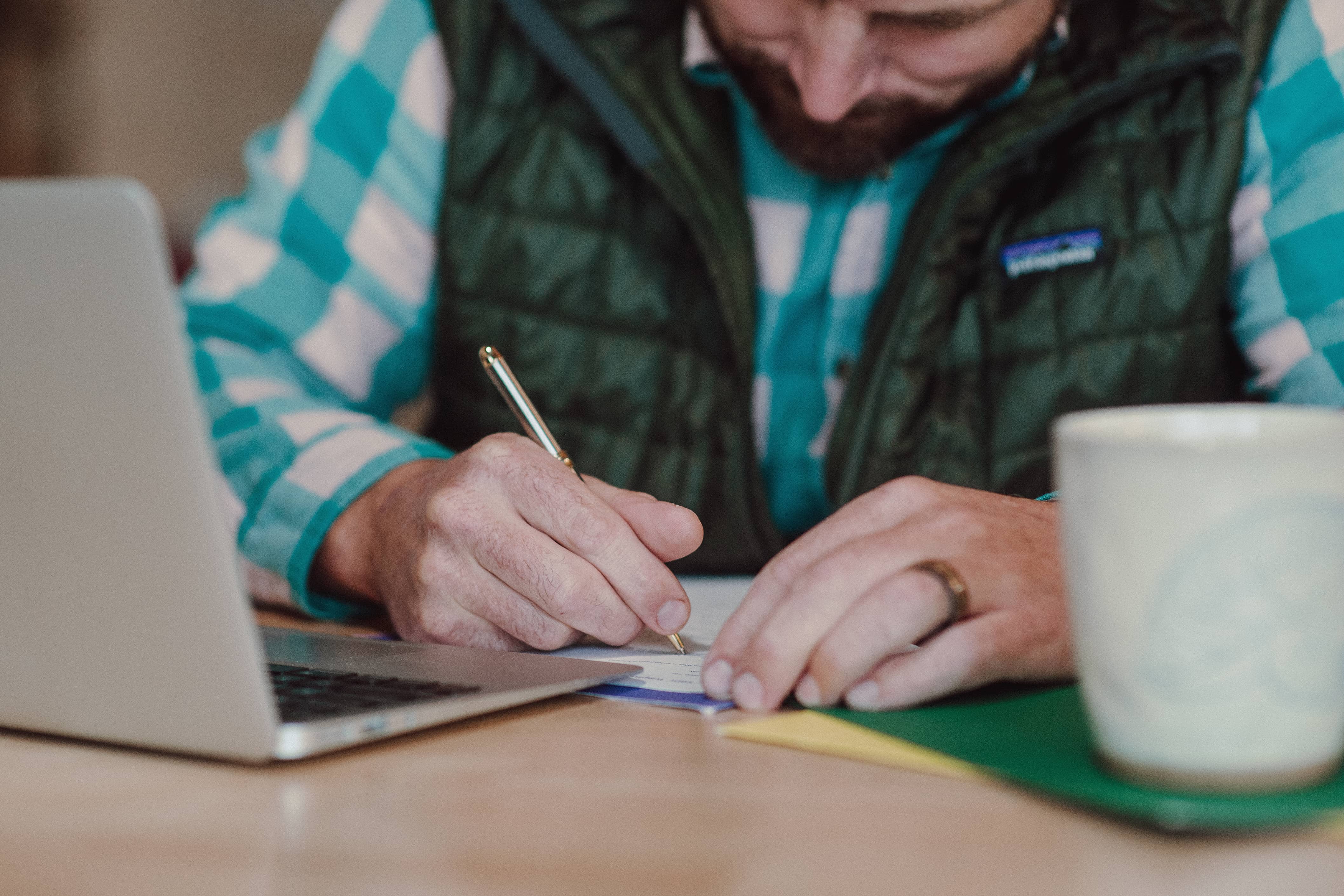 Adult learner writes notes at a table beside a laptop while participating in coursework or educational planning