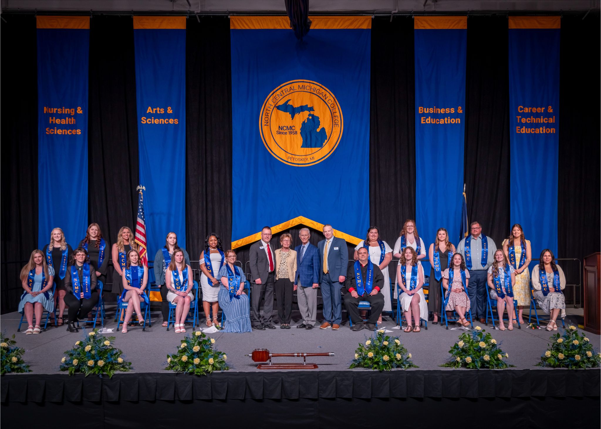 Group photo of North Central Michigan College’s 2025 nursing graduates on stage at the nurse pinning ceremony, joined by Pier and Renee Borra, NCMC President David Roland Finley, and Dean of Nursing, Health Sciences and Public Safety Brent LaFaive. Blue and gold academic banners hang behind the group.