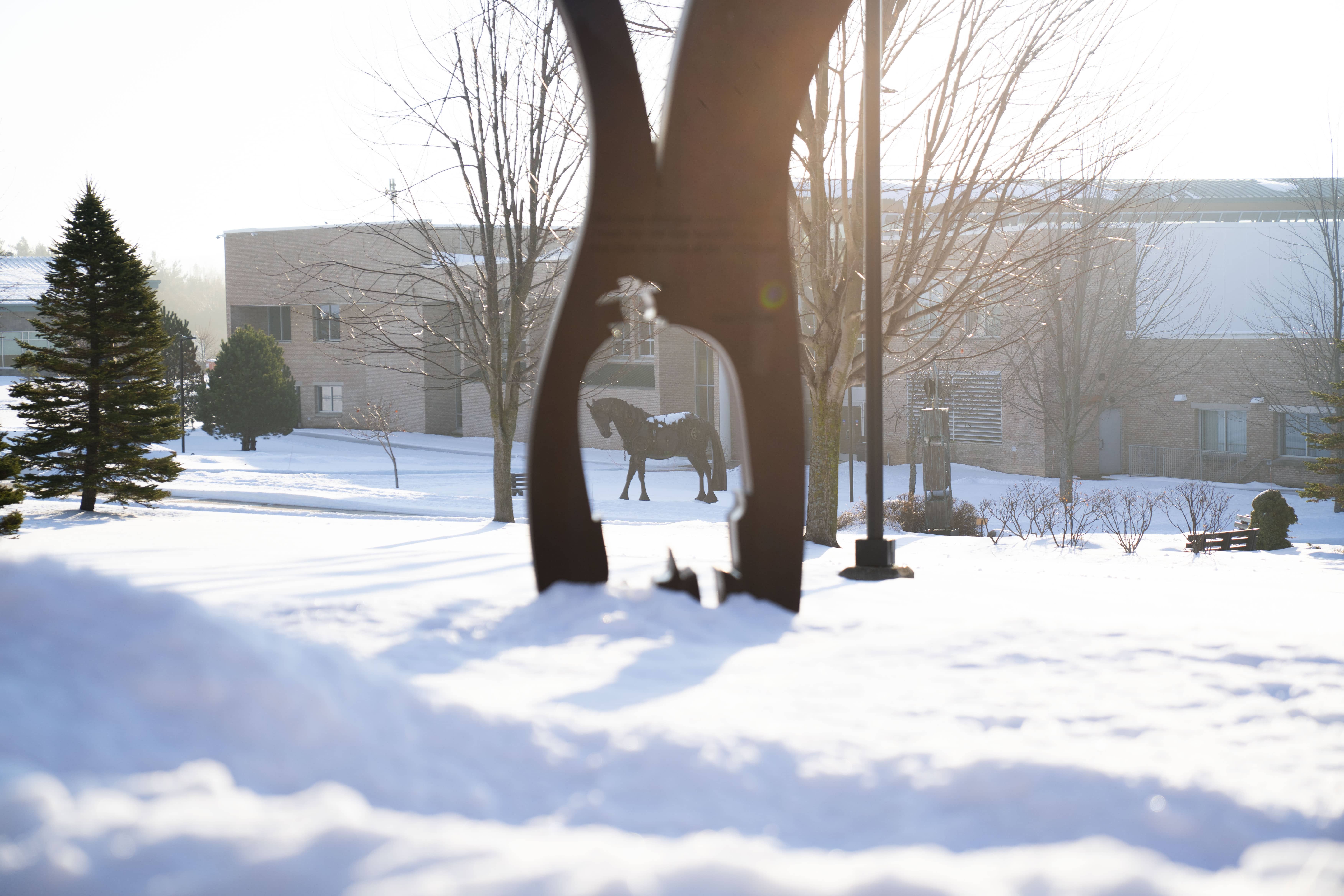 Snow-covered North Central Michigan College campus with a horse sculpture visible through a metal art installation in the foreground